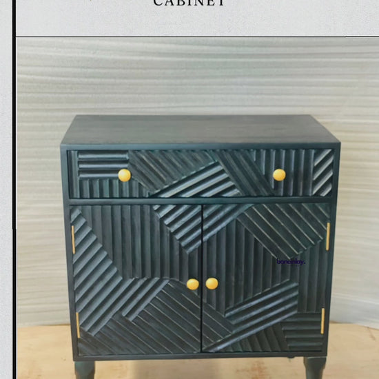 A product video showcasing a hand-carved solid wood cabinet in a deep charcoal or matte indigo finish. The piece features a striking geometric "Paje" design with multi-directional linear carvings across the drawer and doors, accented by minimalist brass-toned knobs. The video demonstrates the functional storage with a smooth-glide drawer and spacious double-door interior, standing on elegant tapered legs.
