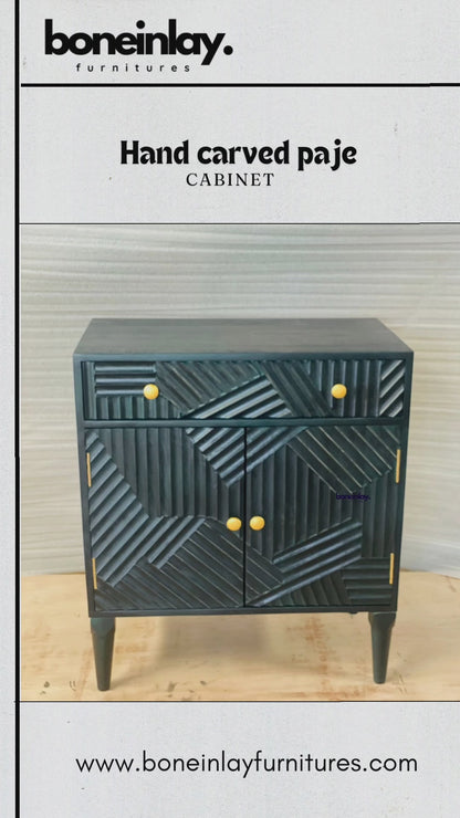 A product video showcasing a hand-carved solid wood cabinet in a deep charcoal or matte indigo finish. The piece features a striking geometric "Paje" design with multi-directional linear carvings across the drawer and doors, accented by minimalist brass-toned knobs. The video demonstrates the functional storage with a smooth-glide drawer and spacious double-door interior, standing on elegant tapered legs.