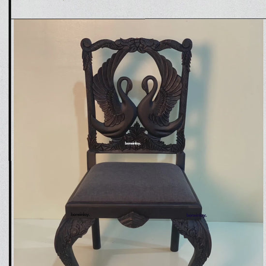 This video features a "Handcarved Menagerie Swan Dining Chair with Arm," highlighting a pair of elegantly hand-carved swans facing each other as the central focus of the backrest. The solid wood frame is finished in a deep espresso tone, while the seat is upholstered in a smooth, dark gray fabric. Close-up shots emphasize the intricate feather details of the swans and the masterfully sculpted cabriole legs, blending artisanal nature-inspired art with functional luxury.
