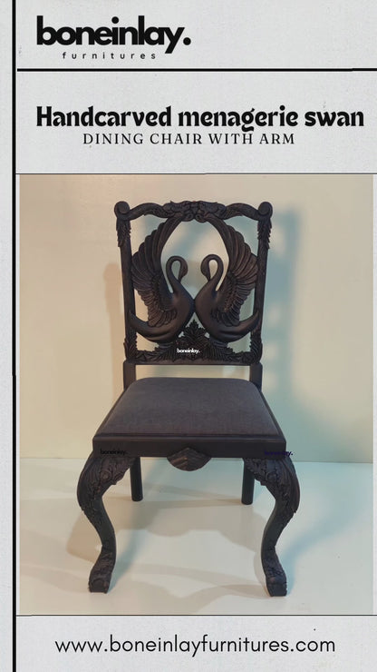 This video features a "Handcarved Menagerie Swan Dining Chair with Arm," highlighting a pair of elegantly hand-carved swans facing each other as the central focus of the backrest. The solid wood frame is finished in a deep espresso tone, while the seat is upholstered in a smooth, dark gray fabric. Close-up shots emphasize the intricate feather details of the swans and the masterfully sculpted cabriole legs, blending artisanal nature-inspired art with functional luxury.