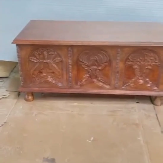 This video showcases a "Hand-Carved Menagerie Coffee Table" in a warm natural wood finish. The camera pans across the detailed front carvings, highlighting the intricate depictions of an owl, a stag, and a central tree of life. A hand opens the lid to reveal a spacious, unfinished wood interior with sturdy gold-toned hinges, demonstrating its dual role as a coffee table and storage trunk. The video concludes with a "Book Now" call to action and highlights including free shipping and customization.