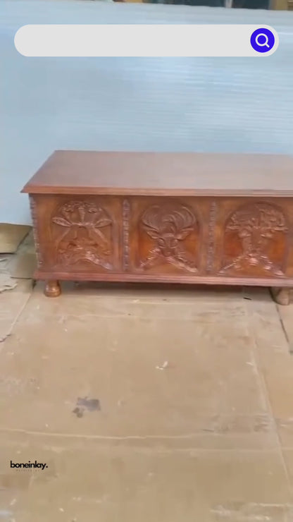 This video showcases a "Hand-Carved Menagerie Coffee Table" in a warm natural wood finish. The camera pans across the detailed front carvings, highlighting the intricate depictions of an owl, a stag, and a central tree of life. A hand opens the lid to reveal a spacious, unfinished wood interior with sturdy gold-toned hinges, demonstrating its dual role as a coffee table and storage trunk. The video concludes with a "Book Now" call to action and highlights including free shipping and customization.