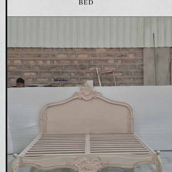 An advertisement video features a beige, handmade hand-carved wooden "Menara Bed" styled in elegant French Rococo design with an intricate shell-and-floral crest headboard, cabriole legs, and detailed relief carvings. The camera pans across the platform frame inside a spacious warehouse, highlighting the subtle color and artisan details from multiple sweeping angles. It concludes with a call-to-action screen displaying "Shop now," the website URL, and consumer incentives including free worldwide delivery an