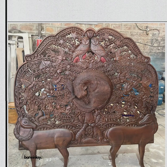 A large, hand-carved wooden headboard from boneinlay furnitures showcasing intricate wildlife motifs set against a rich dark finish. The video pans across detailed carvings featuring a central coiled animal surrounded by foliage, birds, two owls at the top crest, and two prominent deer-like figures forming the base. Shot in a workshop setting, it highlights the artisan craftsmanship, dimensional relief work, and natural wood texture.