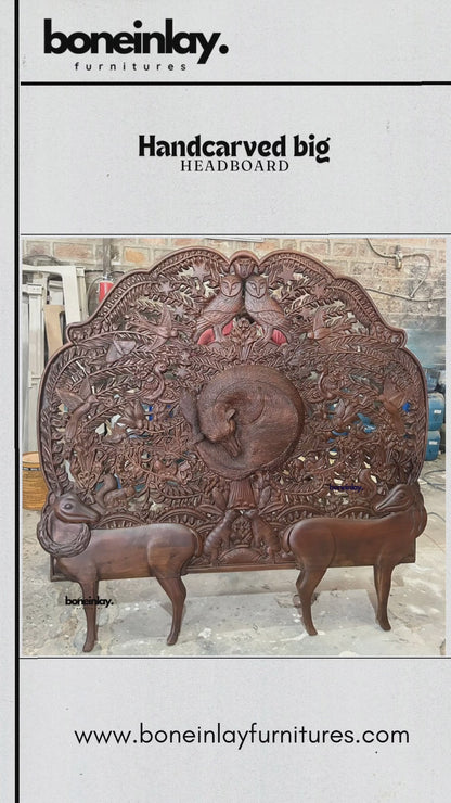 A large, hand-carved wooden headboard from boneinlay furnitures showcasing intricate wildlife motifs set against a rich dark finish. The video pans across detailed carvings featuring a central coiled animal surrounded by foliage, birds, two owls at the top crest, and two prominent deer-like figures forming the base. Shot in a workshop setting, it highlights the artisan craftsmanship, dimensional relief work, and natural wood texture.