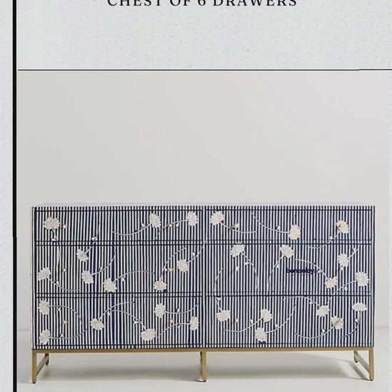 A close-up video showcasing a large, six-drawer indigo blue chest of drawers featuring a striking white bone inlay pattern. The design consists of thin horizontal stripes across the entire piece, accented by delicate floral motifs and round brass knobs. A person is shown opening the drawers, revealing a spacious, dark navy blue interior, emphasizing both the chest’s functionality and its artisanal, textured finish.