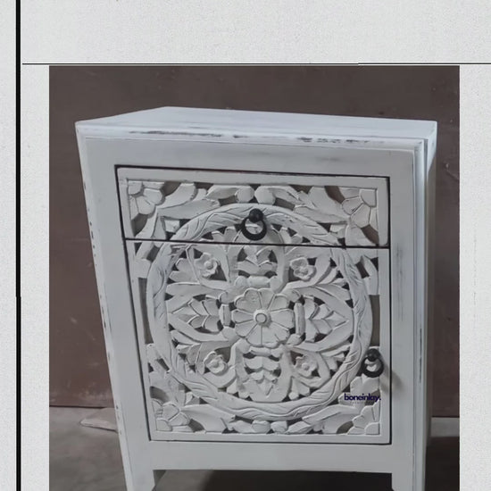 Hand-carved solid wood bedside table with a rustic finish, featuring a circular floral motif across the drawer and cabinet door, dark ring pulls, and shaped legs.
