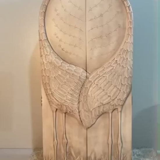 A product video featuring a hand-carved solid wood armoire with a sculptural "Land & Sky" crane motif. The arched doors are intricately carved into the shape of two cranes with their wings meeting in the center, set against a backdrop of delicate branch and leaf patterns. The video showcases the light-wash, distressed artisanal finish and pans to reveal a functional interior equipped with a hanging rod, adjustable shelving, and internal drawers with classic brass hardware.