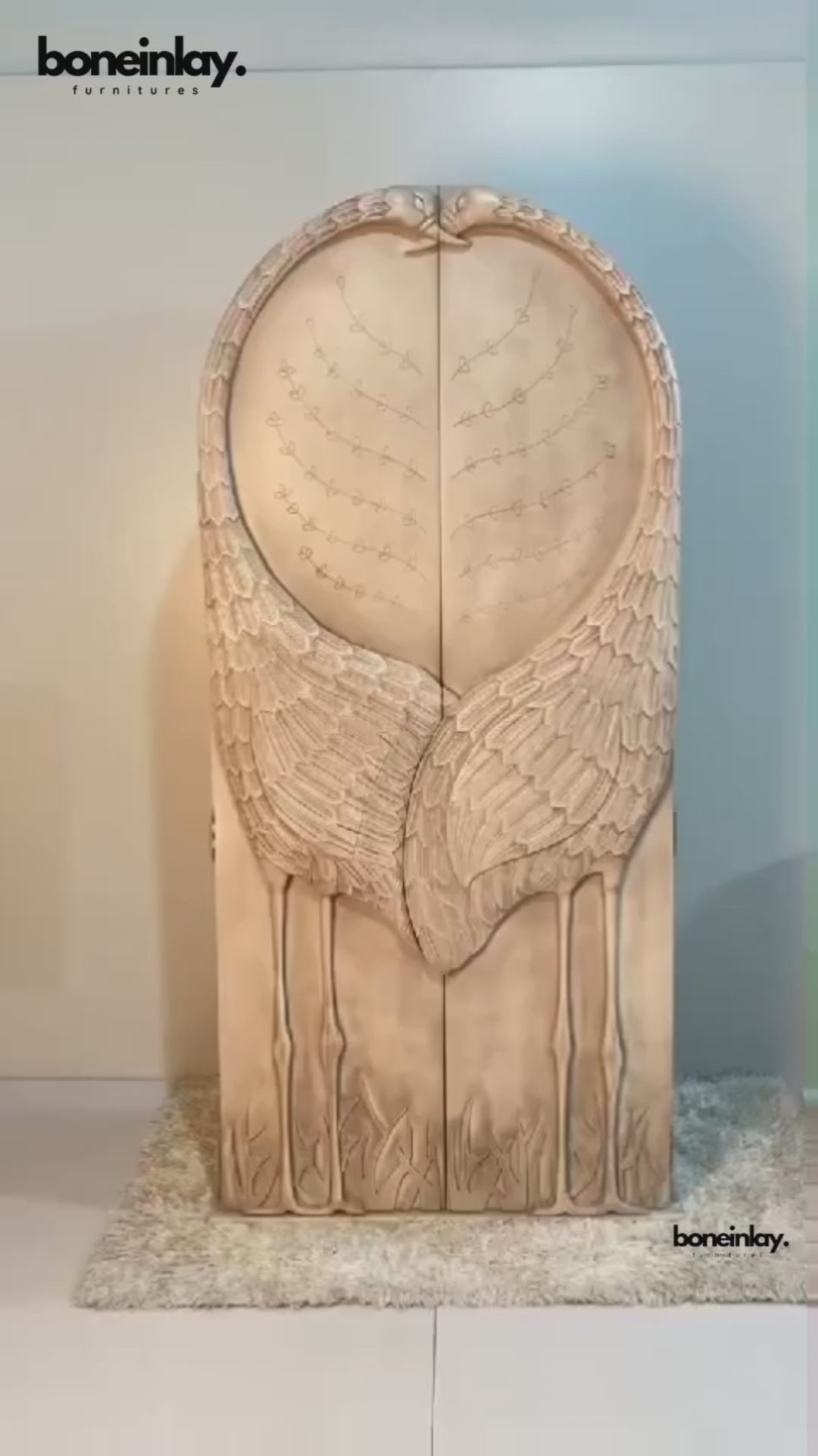 A product video featuring a hand-carved solid wood armoire with a sculptural "Land & Sky" crane motif. The arched doors are intricately carved into the shape of two cranes with their wings meeting in the center, set against a backdrop of delicate branch and leaf patterns. The video showcases the light-wash, distressed artisanal finish and pans to reveal a functional interior equipped with a hanging rod, adjustable shelving, and internal drawers with classic brass hardware.