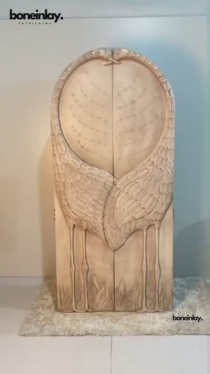 A product video featuring a hand-carved solid wood armoire with a sculptural "Land & Sky" crane motif. The arched doors are intricately carved into the shape of two cranes with their wings meeting in the center, set against a backdrop of delicate branch and leaf patterns. The video showcases the light-wash, distressed artisanal finish and pans to reveal a functional interior equipped with a hanging rod, adjustable shelving, and internal drawers with classic brass hardware.