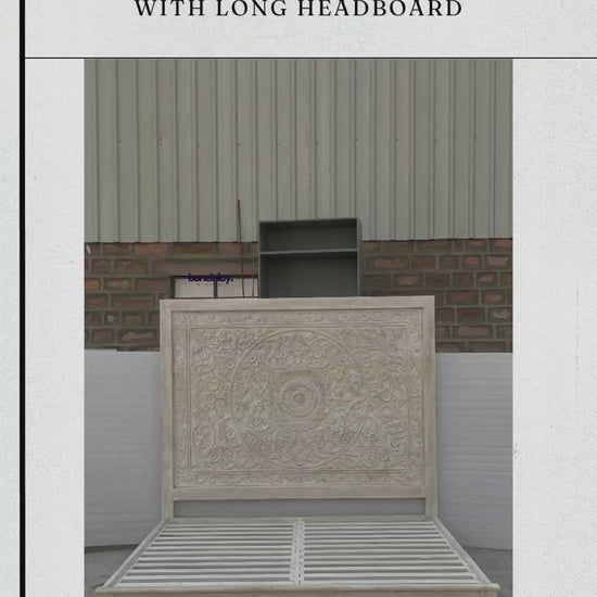A video featuring a rustic hand-carved solid wood bed frame in a light, whitewashed natural finish. The centerpiece is an extra-tall, rectangular headboard featuring a large, intricate circular floral mandala carving at the center, surrounded by detailed vine and leaf motifs. The video highlights the sturdy wooden slat base and the artisanal craftsmanship of the headboard.