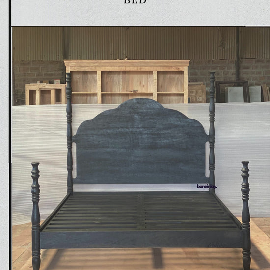 An elegant four-poster bed frame finished in a distressed matte black. The bed features a scalloped, farmhouse-style headboard and intricately turned posts that add a touch of classic craftsmanship. The frame includes a sturdy wooden slat base, blending a bold, moody aesthetic with traditional furniture design.