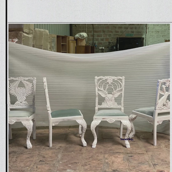 The video showcases intricately hand-carved wooden dining chairs, each featuring a unique animal or nature-inspired motif on the backrest, including an owl, a deer, a tree, and a rabbit. The chairs are painted in a distressed white finish with soft sage green cushioned seats, blending rustic charm with artistic craftsmanship. The scenes highlight the fine detailing of the carvings and the overall elegant design suitable for a menagerie-themed dining space.