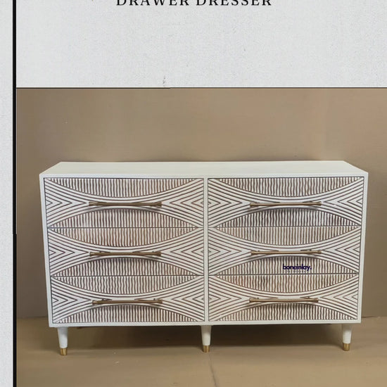 A modern six-drawer white dresser with intricate hand-carved geometric sunburst patterns on each drawer front. The piece features long gold horizontal handles and stands on four white tapered legs with gold metal tips. The high-relief carving creates a textured, two-tone effect between the white surface and natural wood grooves.