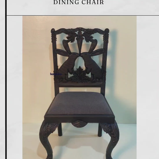 Close-up of a black "Menagerie Bunny" dining chair showcasing detailed hand-carving on the backrest and legs. The chair features a dark charcoal upholstered seat and a whimsical, symmetrical bunny and carrot design carved from solid wood.