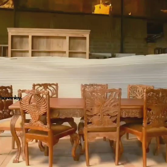 This video showcases an artisan-crafted solid wood dining set with a rich honey-brown finish, featuring an expansive rectangular table and chairs with intricate hand-carved backrests. Each chair displays a unique wildlife or nature motif, including a majestic owl with outspread wings, a peacock, an elephant, and a tree of life, all exhibiting remarkable 3D detail. The set is filmed in an artisanal workshop, emphasizing the traditional wood-carving heritage and the high-quality craftsmanship that makes this 