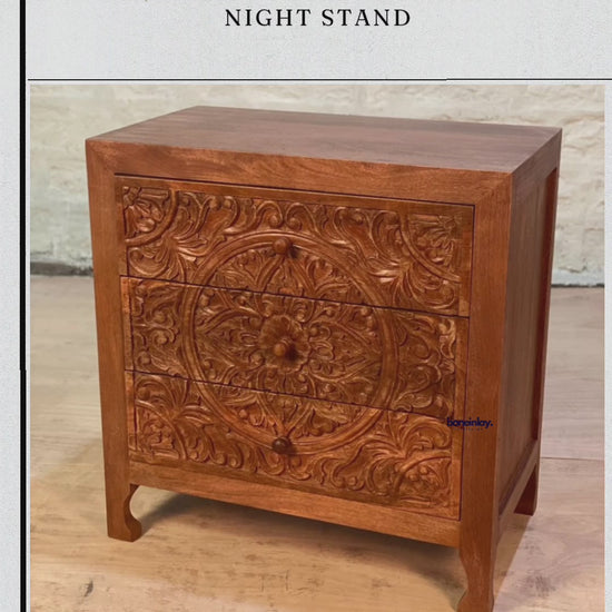A compact, three-drawer wooden nightstand featuring a rich, warm finish and an intricate, hand-carved floral mandala across the drawer fronts. The piece combines a rustic farmhouse feel with elegant, tapered legs and matching round wooden knobs.