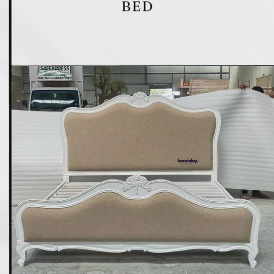 A luxury French Country upholstered bed frame featuring a pristine white hand-carved wooden border and a neutral beige fabric headboard and footboard. The bed displays elegant cabriole legs and ornate floral carvings at the center of the scalloped headboard, set against a workshop backdrop.
