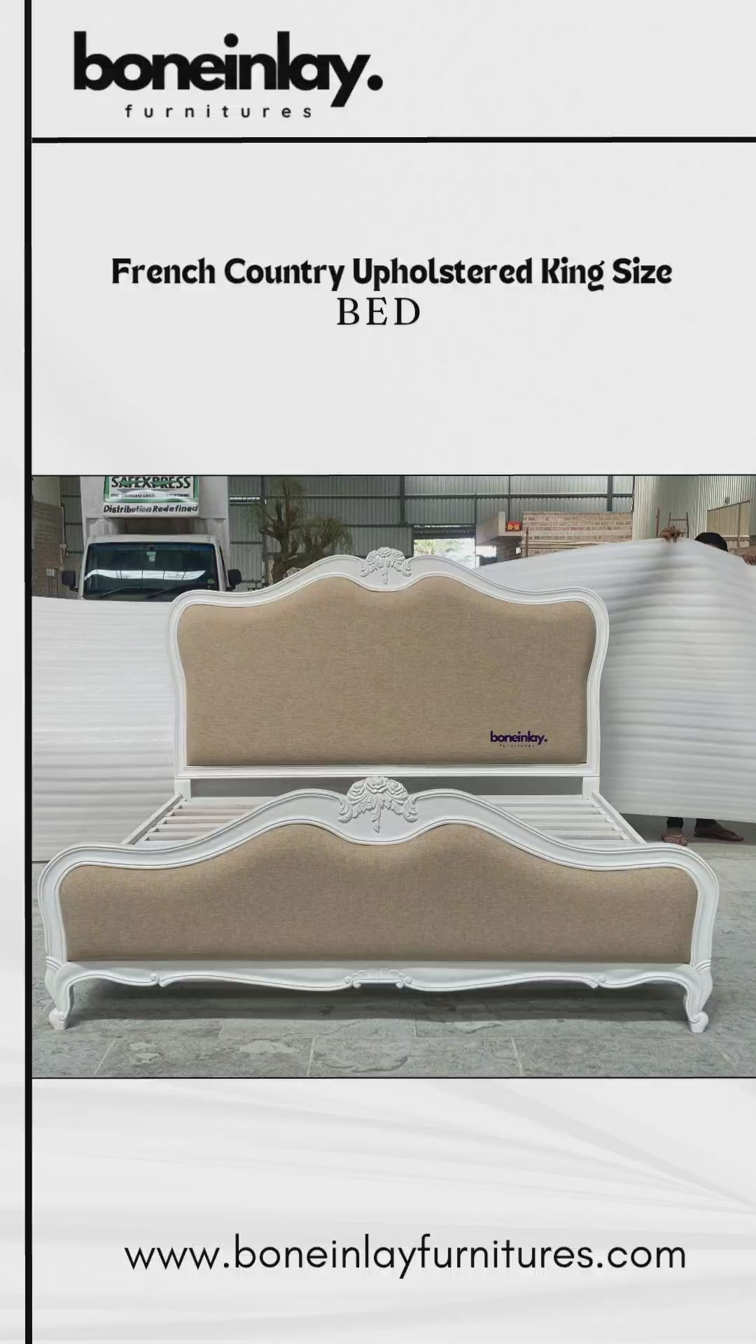 A luxury French Country upholstered bed frame featuring a pristine white hand-carved wooden border and a neutral beige fabric headboard and footboard. The bed displays elegant cabriole legs and ornate floral carvings at the center of the scalloped headboard, set against a workshop backdrop.
