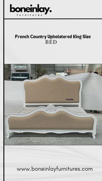 A luxury French Country upholstered bed frame featuring a pristine white hand-carved wooden border and a neutral beige fabric headboard and footboard. The bed displays elegant cabriole legs and ornate floral carvings at the center of the scalloped headboard, set against a workshop backdrop.