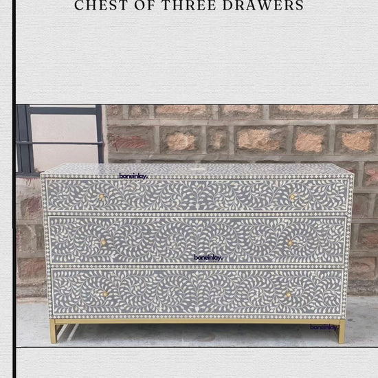 An elegant, grey, handmade chest features an intricate spiral bone inlay pattern of swirling leaves and floral motifs across its three drawers. Each drawer is accented with simple, round gold knobs, complementing the matching gold metal base that elevates the unit. The chest is showcased in an outdoor setting against a rustic brick wall, with a hand opening each drawer to reveal a clean, smooth interior.