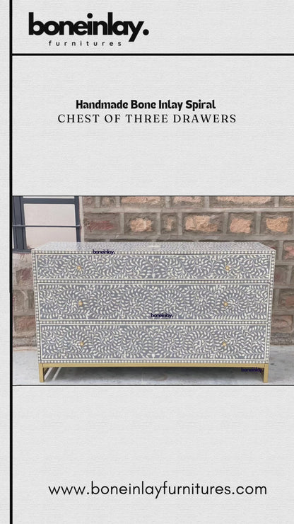 An elegant, grey, handmade chest features an intricate spiral bone inlay pattern of swirling leaves and floral motifs across its three drawers. Each drawer is accented with simple, round gold knobs, complementing the matching gold metal base that elevates the unit. The chest is showcased in an outdoor setting against a rustic brick wall, with a hand opening each drawer to reveal a clean, smooth interior.