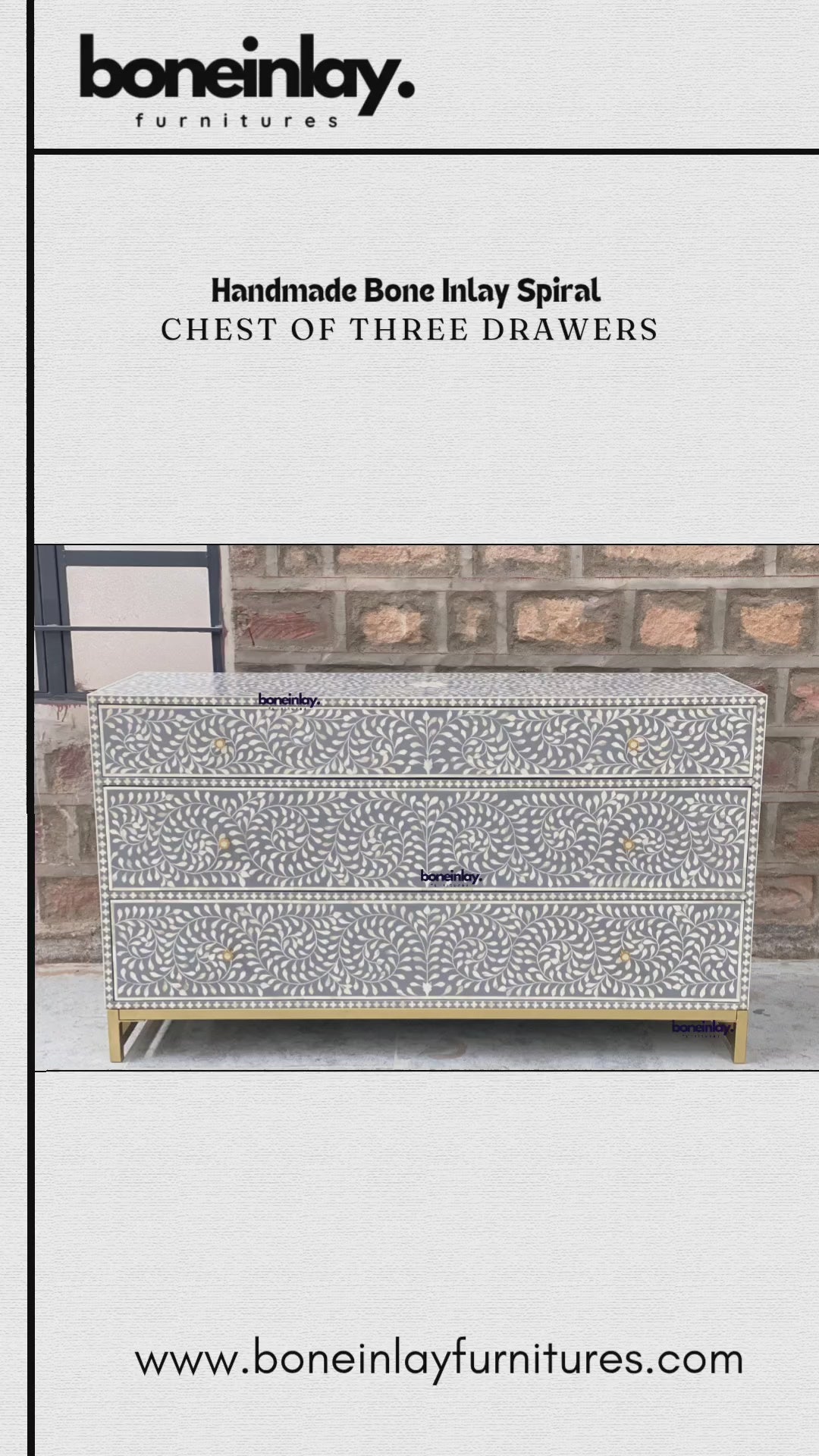 An elegant, grey, handmade chest features an intricate spiral bone inlay pattern of swirling leaves and floral motifs across its three drawers. Each drawer is accented with simple, round gold knobs, complementing the matching gold metal base that elevates the unit. The chest is showcased in an outdoor setting against a rustic brick wall, with a hand opening each drawer to reveal a clean, smooth interior.