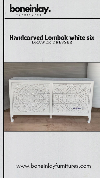 A wide, white-finished wooden dresser featuring six drawers entirely covered in intricate, hand-carved floral and vine motifs. The piece blends traditional craftsmanship with a clean, bohemian aesthetic, supported by four tapered legs. The video highlights the deep texture of the carvings and the smooth, matte surface of the dresser's top.