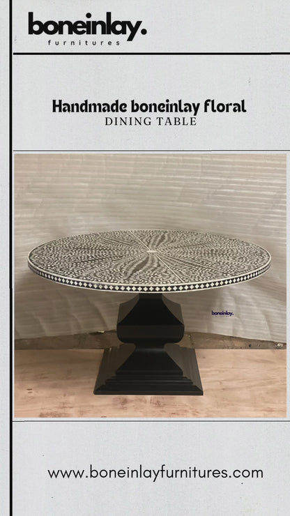A round, handmade bone inlay dining table featuring an intricate black and white floral and leafy pattern that radiates outwards from the center. The ornate, circular tabletop sits atop a sturdy, geometric pedestal base finished in solid matte black. The rim of the tabletop is detailed with a contrasting black and white checkerboard border.