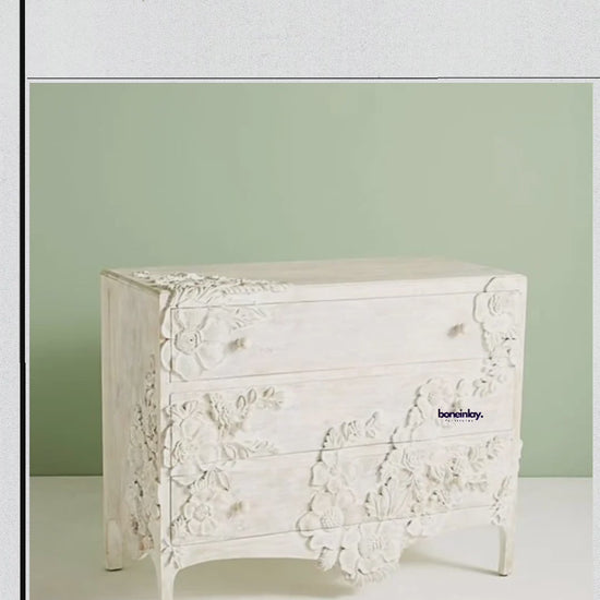 Close-up video of a hand-carved Enchantment Three Drawer Dresser in a whitewashed finish, showcasing intricate 3D floral carvings across the drawer fronts and sides. The video highlights the artisanal craftsmanship and smooth gliding drawers of this vintage-inspired solid wood furniture.