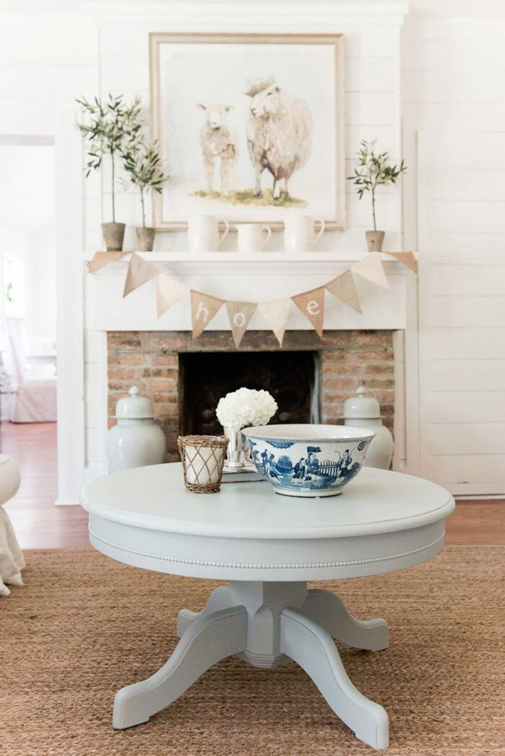 Round wooden pedestal coffee table with curved feet on a woven rug in a cottage-style living room with a fireplace.