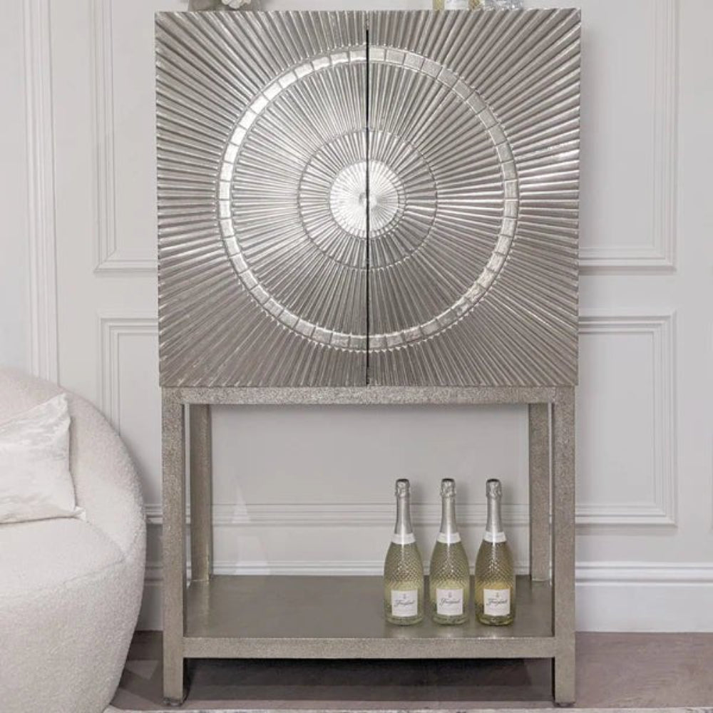 Antique silver hand-embossed bar cabinet with a sunburst radial pattern on two doors and a tall open metal base holding bottles.