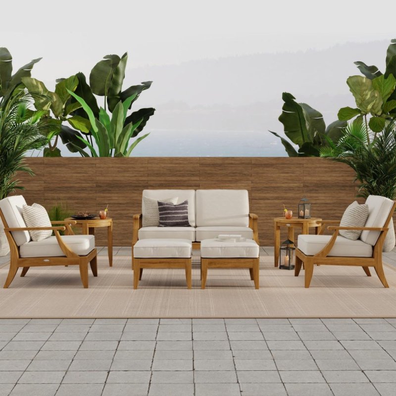 A seven-piece solid wood patio furniture set featuring a cream-cushioned loveseat, two club chairs, ottomans, and round side tables on a modern terrace with tropical plants.