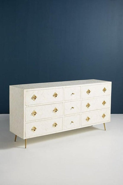 Full front view of a white optical bone inlay 9-drawer dresser with diamond brass handles and gold tapered legs, styled against a deep navy blue wall in a modern bedroom.
