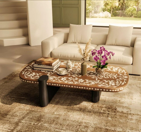An oval-shaped wood coffee table with white floral bone inlay patterns and thick black legs, styled with books and an orchid in a modern living room.