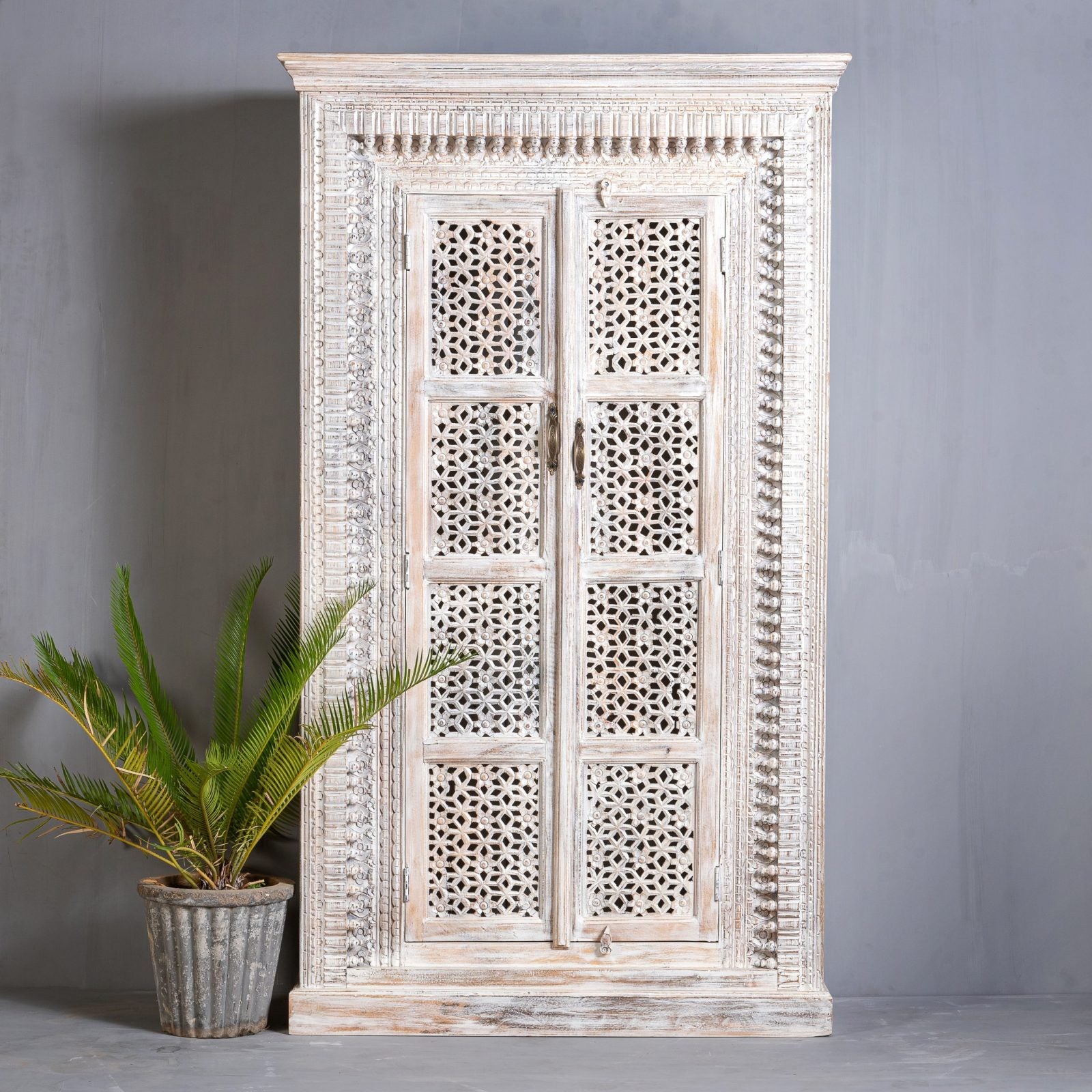 A tall, hand-carved wooden armoire with intricate geometric jali lattice patterns on two doors and a weathered white-wash finish, standing against a grey wall.