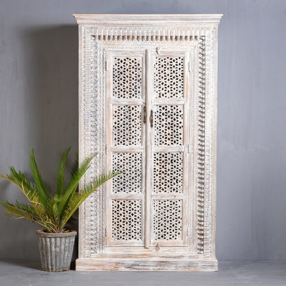 A tall, hand-carved wooden armoire with intricate geometric jali lattice patterns on two doors and a weathered white-wash finish, standing against a grey wall.