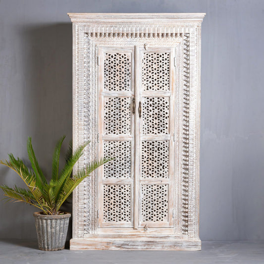 A tall, hand-carved wooden armoire with intricate geometric jali lattice patterns on two doors and a weathered white-wash finish, standing against a grey wall.
