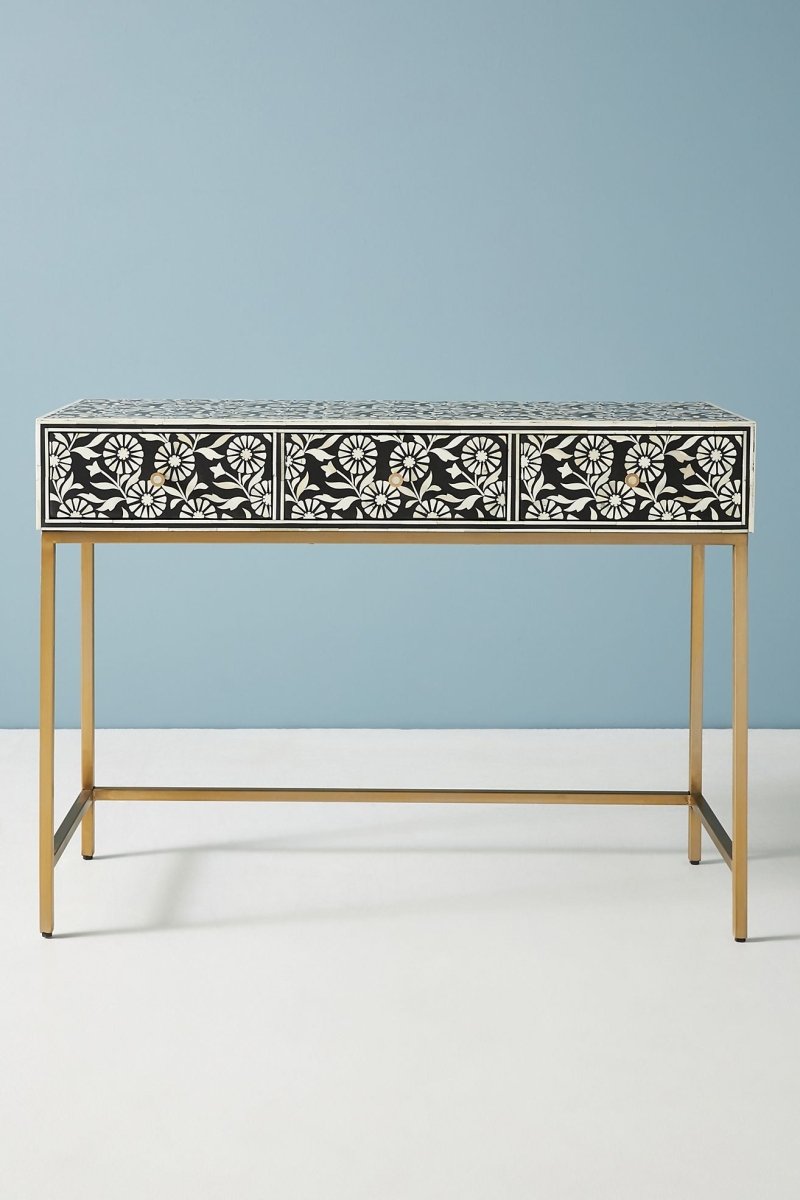 Black and white bone inlay sofa table in a straight front view, showing the full rectangular console with three floral-pattern drawers, brass knobs, a dark top surface, and a brushed gold-toned metal frame base against a soft blue wall.