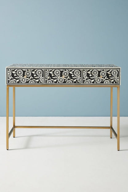 Black and white bone inlay sofa table in a straight front view, showing the full rectangular console with three floral-pattern drawers, brass knobs, a dark top surface, and a brushed gold-toned metal frame base against a soft blue wall.