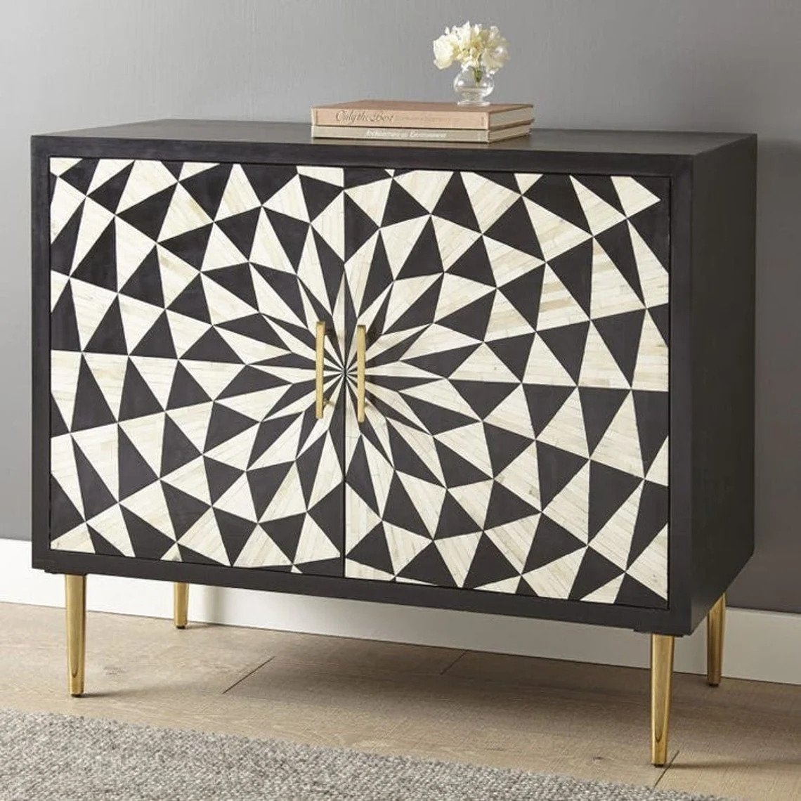 Two-door black and white geometric bone inlay storage cabinet with brass handles and tapered gold metal legs on wooden floor