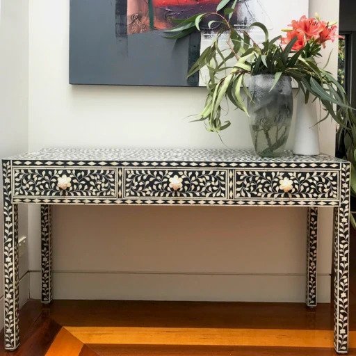 Black bone inlay 3-drawer console table with floral vine inlay and bone knobs, styled in a warm home entryway with timber flooring and floral arrangement.