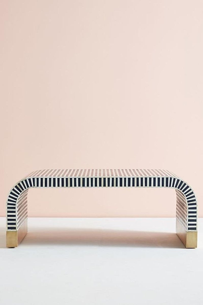 Front angle view of a handmade navy and white bone inlay waterfall coffee table with a rounded rectangular top, flowing striped inlay over the curved sides, and gold-tone block bases against a pale studio background. The open base shows the table's long low profile.
