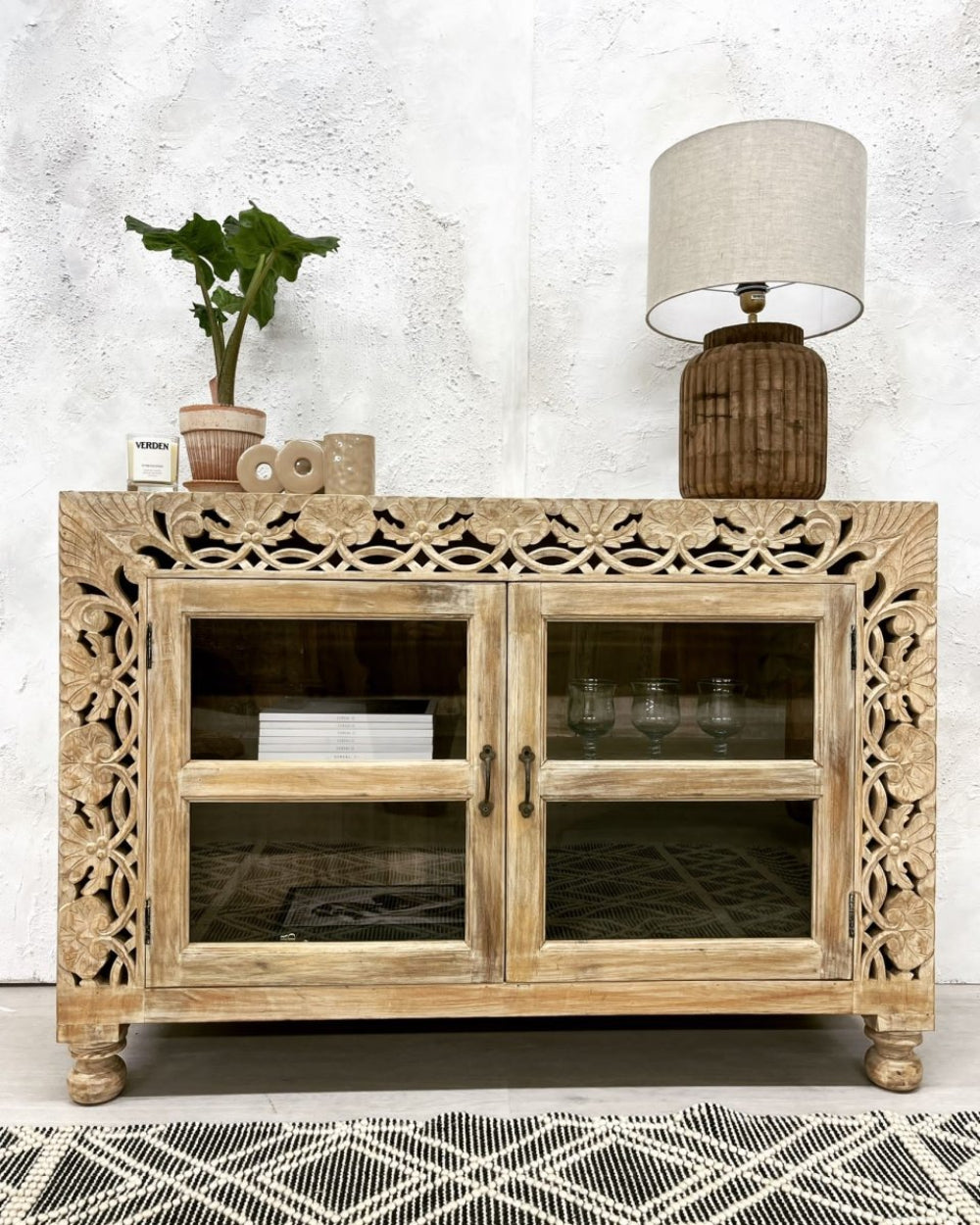 Floral carved wood sideboard in a rustic finish, front view with glass doors, turned feet and a lamp on top.