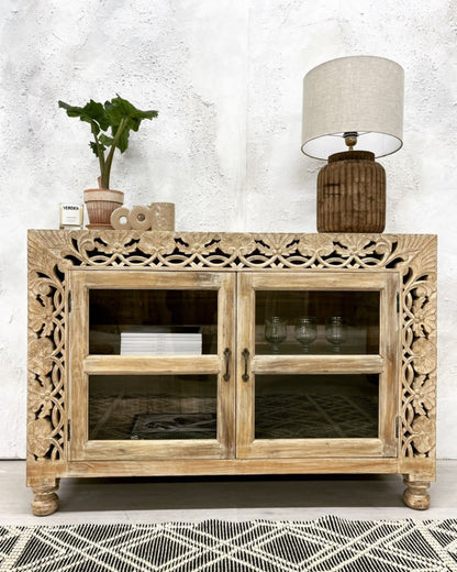 Floral carved wood sideboard in a rustic finish, front view with glass doors, turned feet and a lamp on top.