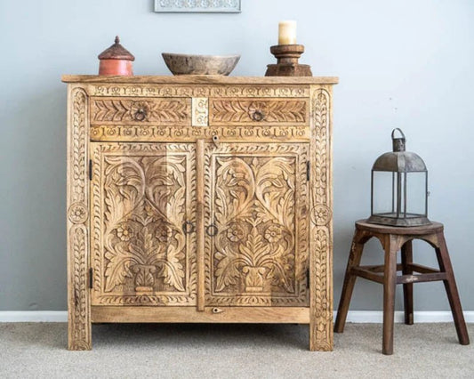 Floral Wood carved cabinet in natural wood, front view showing carved double doors and ring pulls beside a lantern-topped stool.