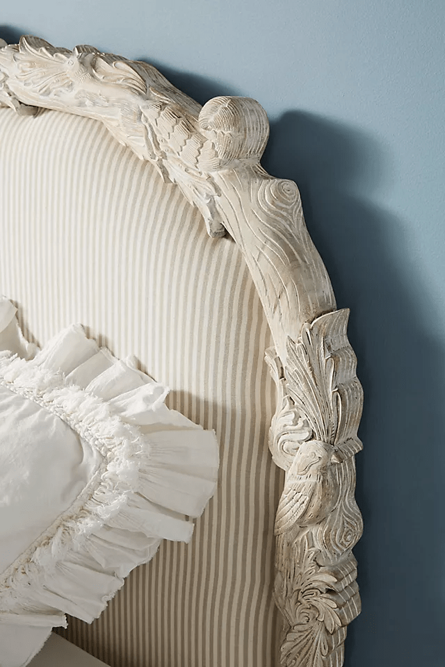 Detailed corner of the Menagerie headboard in whitewashed mango wood,
showcasing hand-carved acanthus leaves, scrollwork, and turned finial,
framing neutral pinstripe linen upholstery with a ruffled white pillow.