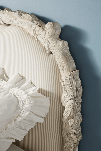 Detailed corner of the Menagerie headboard in whitewashed mango wood,
showcasing hand-carved acanthus leaves, scrollwork, and turned finial,
framing neutral pinstripe linen upholstery with a ruffled white pillow.