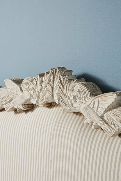 Macro detail of the Menagerie headboard crest in whitewashed mango wood,
featuring hand-carved hummingbirds, acanthus scrolls, and floral motifs,
set against neutral pinstripe linen upholstery fabric.