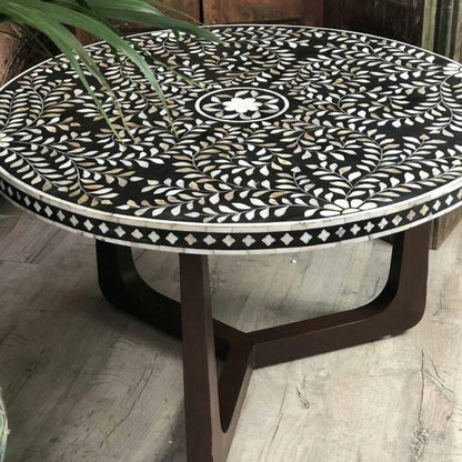 Handcrafted black floral bone inlay round coffee table shown in a front angle, with a black-and-white floral vine pattern across the tabletop and brown legs supporting the rounded solid-wood base for a living room or hallway setting.
